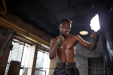 Fototapeta na wymiar Muscular shirtless African boxer fighting with punching bag during sports training in gym