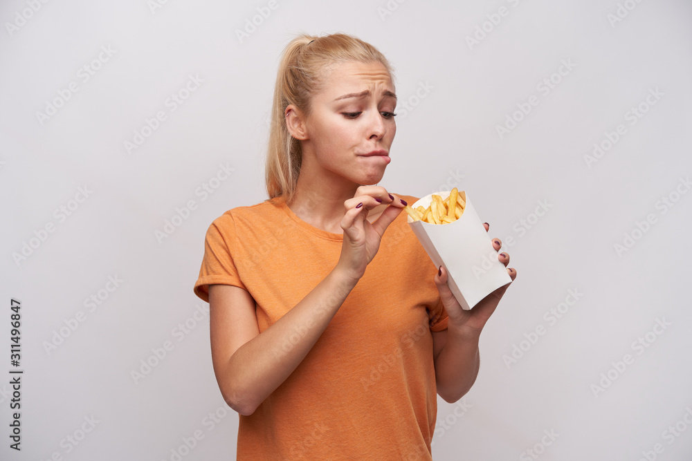 Wall mural Indoor shot of young long haired blonde lady keeping paper box with french fries and looking excitedly at it, wanting to eat it, but worrying about extra calories, isolated over white background