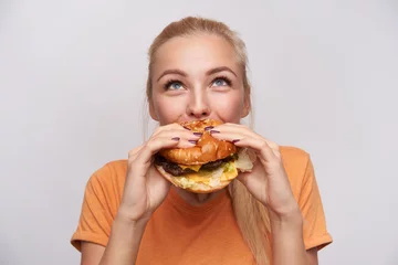 Zelfklevend Fotobehang Portrait of pleased young lovely blonde woman with casual hairstyle eating fresh hamburger with great appetite and looking cheerfully upwards, posing over white background © timtimphoto