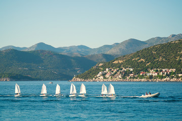 A line of small sailing boats with kid sailors on a tugboat is passing by the hilly seashore