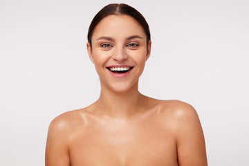 Cheerful attractive young dark haired lady with natural makeup wearing ponytail hairstyle while standing against white background, laughing happily while looking at camera