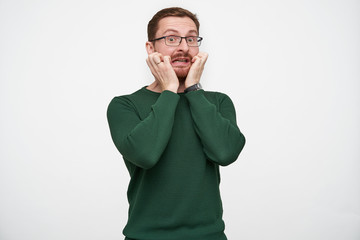 Indoor shot of scared young brunette bearded man in eyewear holding raised hands on his cheeks and crooking face afraid, standing against white background in casual clothes