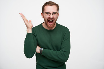 Portrait of angry young bearded guy in eyewear with brown short hair raising affectively his hand and screaming fiercely with wide mouth opened, isolated over white background