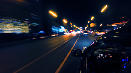 A dark-colored car is moving rapidly along the illuminated street of a night city, on a blurred...