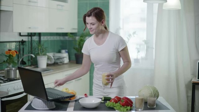 online recipe, young woman measures a wheat porridge with spoon during making salad and watching laptop to screen at the table in the kitchen