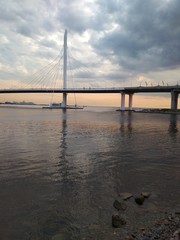 View of the beautiful landscape. Beautiful sky with dark clouds and sunset. An interesting cable-stayed bridge, below a wide river, in which there are stones in the foreground