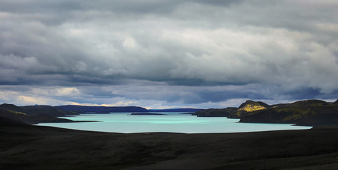 türkisfarbener See in isländischer Lavawüste mit Sonnenstrahl durch Wolkendecke