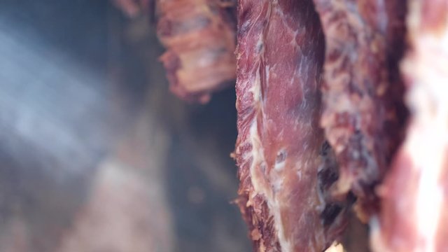 Hanging bacon in smokehouse on blue smoke in slow motion. Piercing sun rays. Drying meat in the traditional way.