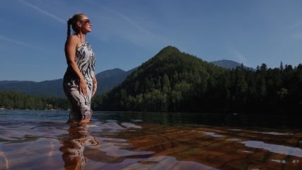 Woman walk on water on pier in sunglasses and a boho silk shawl. Girl rest on a flood wood underwater dock. The pavement is covered with water in the lake. In the background are mountain and a forest.
