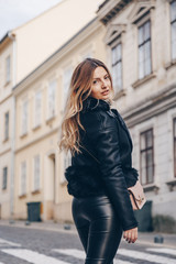 Plakat street style portrait of attractive blonde woman wearing leather trousers and jacket with a trendy purse. crossing the street and smiling at camera. fashion outfit perfect for autumn fall winter