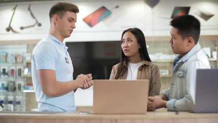 Young Asian couple choosing laptop computers at gadget store and asking questions to male shop assistant