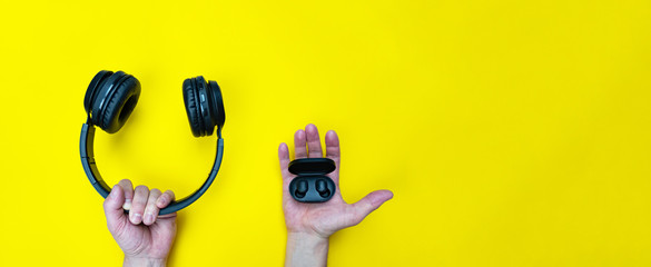 Small black wireless earphones and big headphones in mans hands on yellow background. What to...
