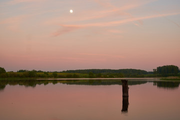 sunset on the shore of a pond with calm water, three cell towers and the moon in the sky.
