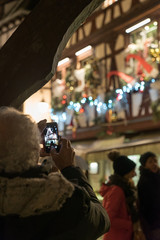 senior citizen taking a photo with cell phone  of beautifully decorated half-timbered houses in La Petite France in historic Strasbourg during the traditional Christmas market