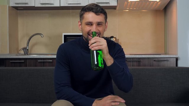 A young man in dark blue pullover and gray pants sitting on the couch, drinking beer, laughs and watches entertainment on TV. Kitchen on background. 4k slowmotion footage