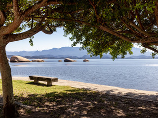 Fototapeta na wymiar Tree, shadow, concrete bench, sea and mountains, Catimbaú Beach, Paqueta, Rio de Janeiro, Brazil