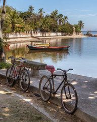 Fototapeta premium Two bicycles standing in the shade, with boat, sea and trees in the background, blue sky, Paqueta Island, Rio de Janeiro, Brazil