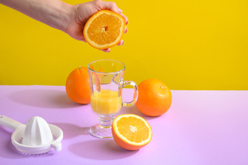 Naklejka premium Juicy fresh orange in female hands hovers over a glass. Against a bright background, purple-yellow. Hard creative light, horizontal photo