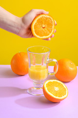 Juicy fresh orange in female hands hovers over a glass. Against a bright background, purple-yellow