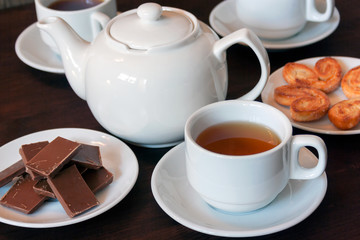 White porcelain teapot, tea cups with tea and sweets on a dark wooden table.
