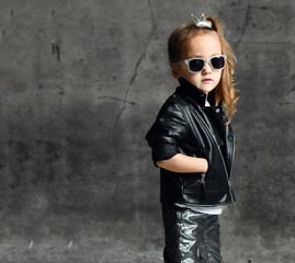 Obraz premium Cool brutal kid girl in leather jacket, jeans and sunglasses is posing like a biker rockstar on concrete wall background