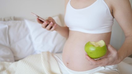 Closeup Of Pregnant Woman Using Smartphone And Eating Green Apple.