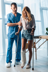 handsome doctor looking at injured woman walking with crutches