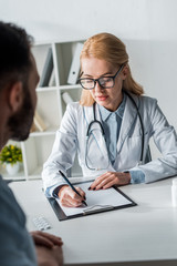 selective focus of attractive doctor in glasses writing prescription near man