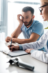 selective focus of shocked bearded man looking at doctor writing diagnosis