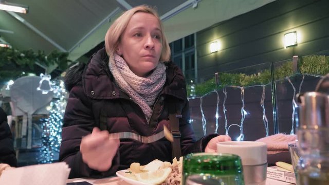 Middle Aged Woman Eating Chips With A Fork In A Diner At Night. Outdoors Restaurant Terrace At Christmas Winter Time.