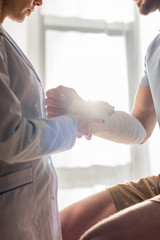cropped view of orthopedist touching injured hand of man in clinic