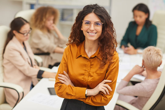 Elegant woman with long curly hair standing with arms closed wearing eyeglasses looking at camera smiling with colleagues coworking behind her, medium portrait