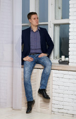 Young man with an arrogant expression. Sits by the window on the windowsill.