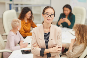 Attractive Asian woman standing with arms crossed looking at camera with colleagues coworking behind her, waist up portrait