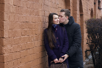 Fototapeta na wymiar A young man kisses a girl on the cheek. Against the background of a brown brick wall. Dressed in a coat and jacket.