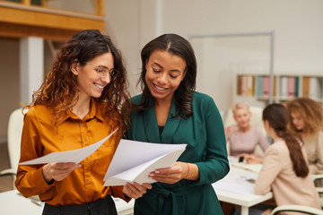 Two attractive stylish women choosing best design sketch, their colleagues coworking behind them