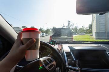 Close up young man car driver drink coffee, hand holding a Paper Cup of coffee in the background steering the car dashboard blurry green background.