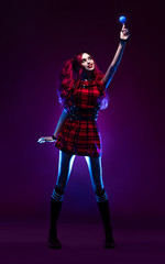 Attractive woman with red hair in checkered dress holding blue lollipop and posing on dark background