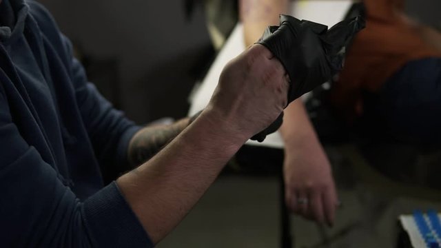 Close up of hands tattoo artist putting on the latex black gloves before a tattoo session at the tattoo shop. Tattooist sitting on chair in front the unrecognizable client. Slow motion