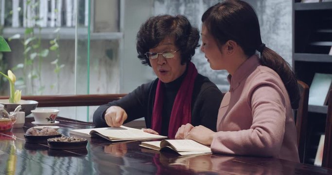 Chinese senior profesor teaching young grace woman traditional literature reading book together, senior mentor instructing student Chinese culture discussing talking together at home or library