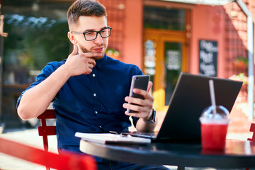 Thoughtful young businessman holding modern smartphone. Freelancer with a mobile phone thinks about reply. Man in glasses with laptop on table in outdoor cafe. Multitasking and telecommuting theme.