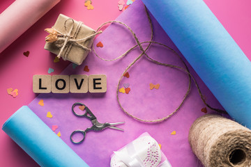 top view of valentines decoration, scissors, wrapping paper, twine, gift boxes and love lettering on wooden cubes on pink background