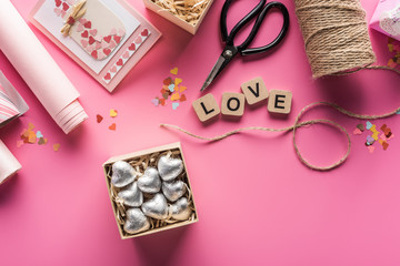 top view of valentines decoration, scissors, wrapping paper, twine, gift box with silver hearts, greeting card and love lettering on wooden cubes on pink background