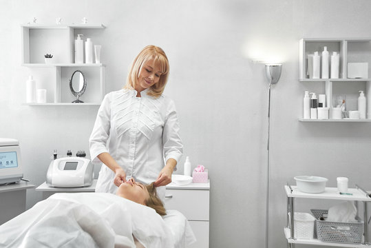 Professional beautician gives young woman manual massage of face and cervical collar zone in white beauty parlor. Сosmetology, face care procedure