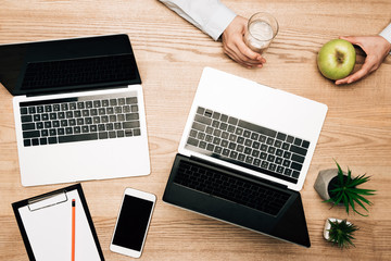 Top view of businessman holding apple and glass of water by laptops and clipboard on table