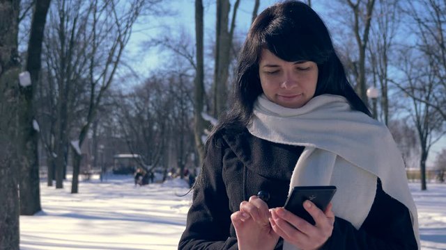 Happy Smiling Beautiful Girl Using Smart Phone. Sunny Day In Snow Covered City Park. Slow Motion 30 fps 0.5 normal speed 60 fps