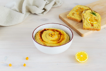 Hummus in a bowl with sandwiches on a light wooden table.