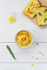 Hummus in a bowl with sandwiches on a light wooden table.