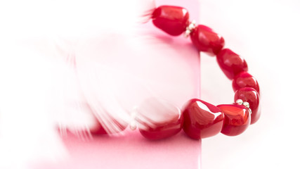 Glass jewellery or blood coral red coloured bijou or gemstone wristband or necklace on pale pink gift box with elegant blurred white feather background. Selective focus. Abstract love gift background.