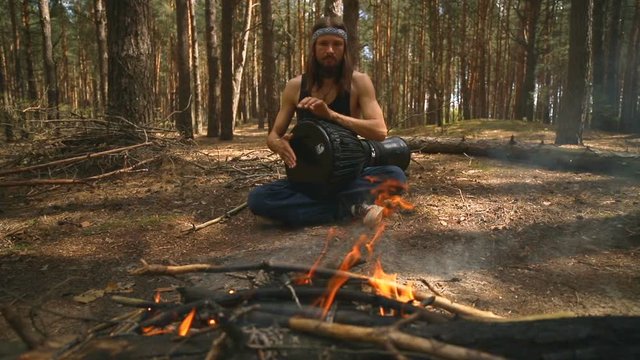 Young Caucasian hippie man playing a djembe drum in the summer coniferous forest